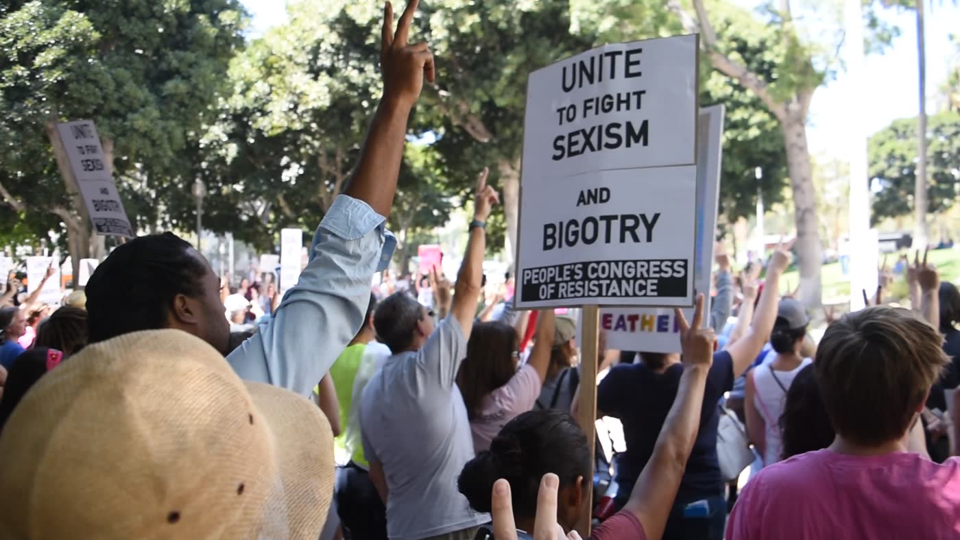 a rally crowd in downtown los angeles, hands raised, a sign reading unite to fight sexism and bigotry